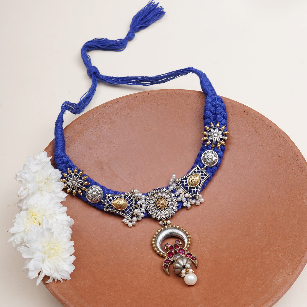 A royal blue braided necklace with ornate silver-toned and gold-toned metal embellishments, white pearl-like beads, and a dangling decorative pendant.